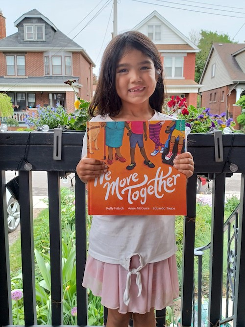 A photo of Aisha. She is 6 years old and has long, straight black hair. She is wearing a pink skirt and standing on a porch, grinning and holding a copy of the book
