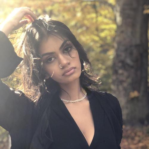 A photo of Ahona Mehdi. Ahona has shoulder-length black hair, brown skin, and they are wearing a black shirt and a nose ring. She is standing in a wooded area with sunlight filtering through the leaves.