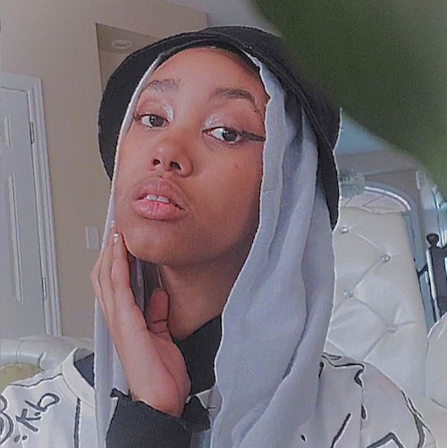 A photo of Amani Omar. Amani has brown skin and is wearing black winged eyeliner, a light blue hijab, and a black bucket hat on top. She is looking sideways at the camera with her hand resting on her chin.