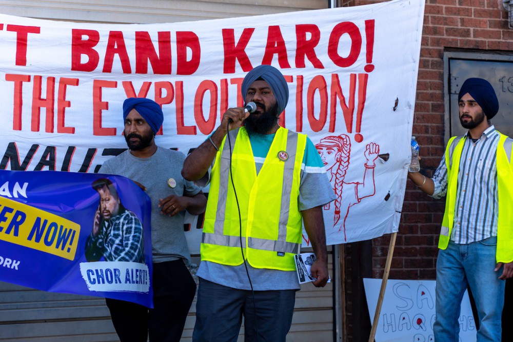 Attar, a tall man with dark brown skin and a blue turban, wears a high-viz vest and speaks into a microphone. Behind him are two turbaned people holding banners.