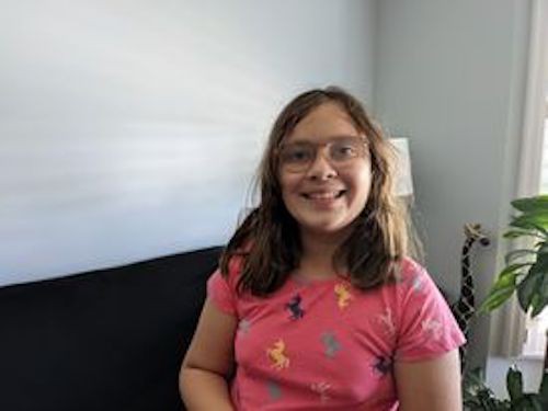 A photo of Divya. Divya is 9 years old, has long brown hair and glasses, is wearing a pink T-shirt, is sitting on a couch, and smiling at the camera.