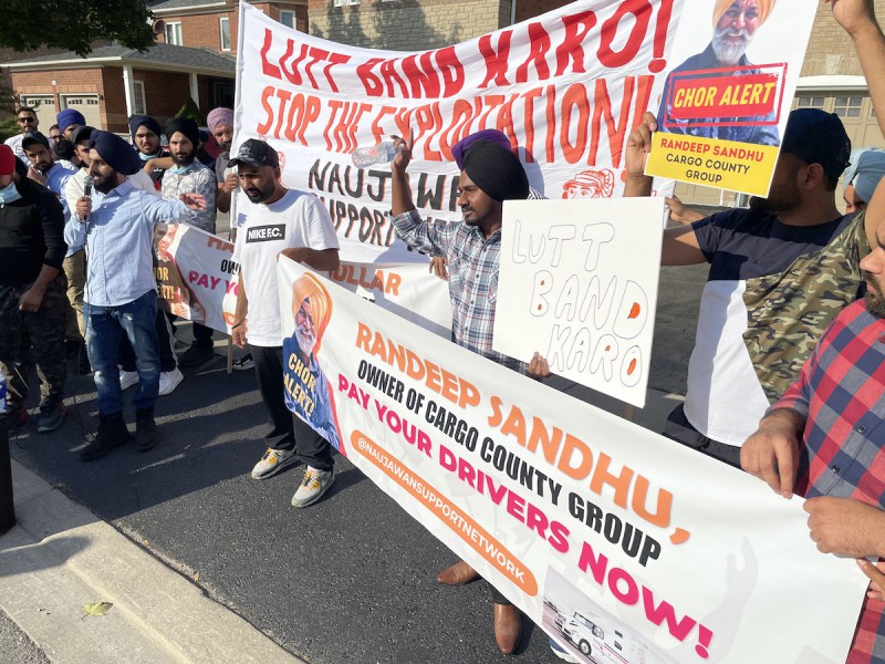 A photo of a crowd of brown men – some wearing turbans, some wearing baseball caps – holding a banner that says "Randeep Sandhu, owner of Cargo County Group: Pay your drivers now!" They are standing in front of a residential house, and one is speaking into a microphone.