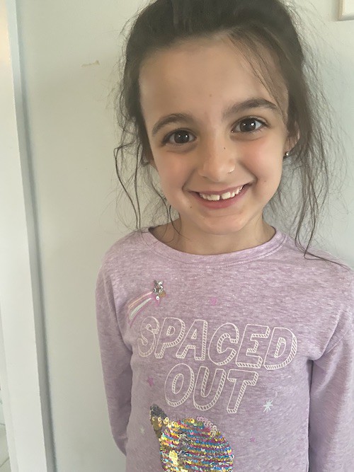 A photo of Isabelle. She is 6 years old and has brown hair pulled back into a messy ponytail. She is wearing a purple shirt with the words "SPACED OUT" and is grinning at the camera.