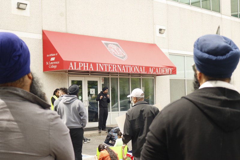 A photo of Jaspreet standing under a red awning that reads "Alpha International Academy." He wears a black turban and is speaking into a microphone. The photo is taken from within a crowd watching Jaspreet.