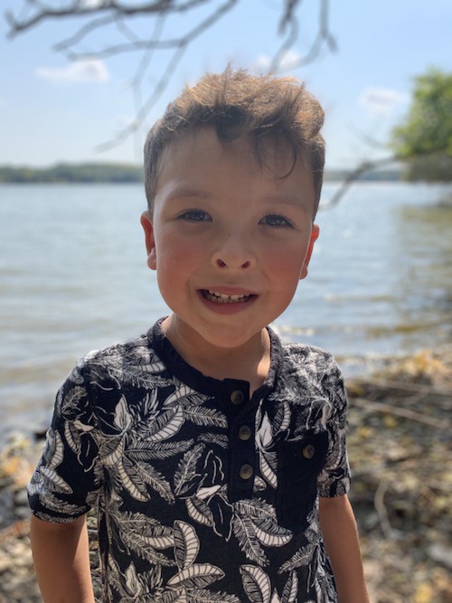 A photo of Langston. He is 6 years old, and has short, curly brown hair. He's standing on a sunny beach and wearing a navy-and-white shirt.