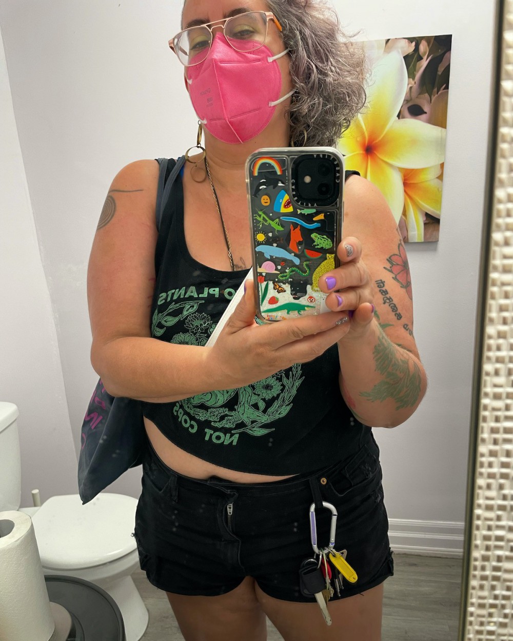 A mirror selfie of Leah, a 47 year old nonbinary femme with light brown skin, curly silver, lavender and brown hair on one side of their head, rose gold aviator glasses and a hot pink N95 mask, wearing a navy and turquoise crop top that says