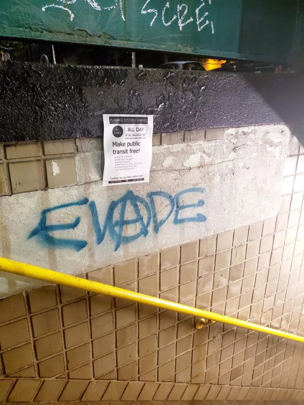 Graffiti in a stairway of the NYC subway that says "EVADE." The A is surrounded by a circle. Above it, a poster says "Planned system changes: Make public transit free!"