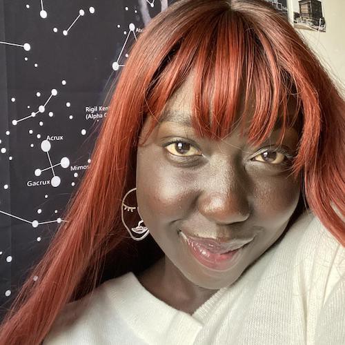 A photo of Mary Cep. Mary has dark skin and long, straight, reddish-brown hair with bangs. She is wearing a silver earring in the shape of a face, and looking at the camera and smiling.