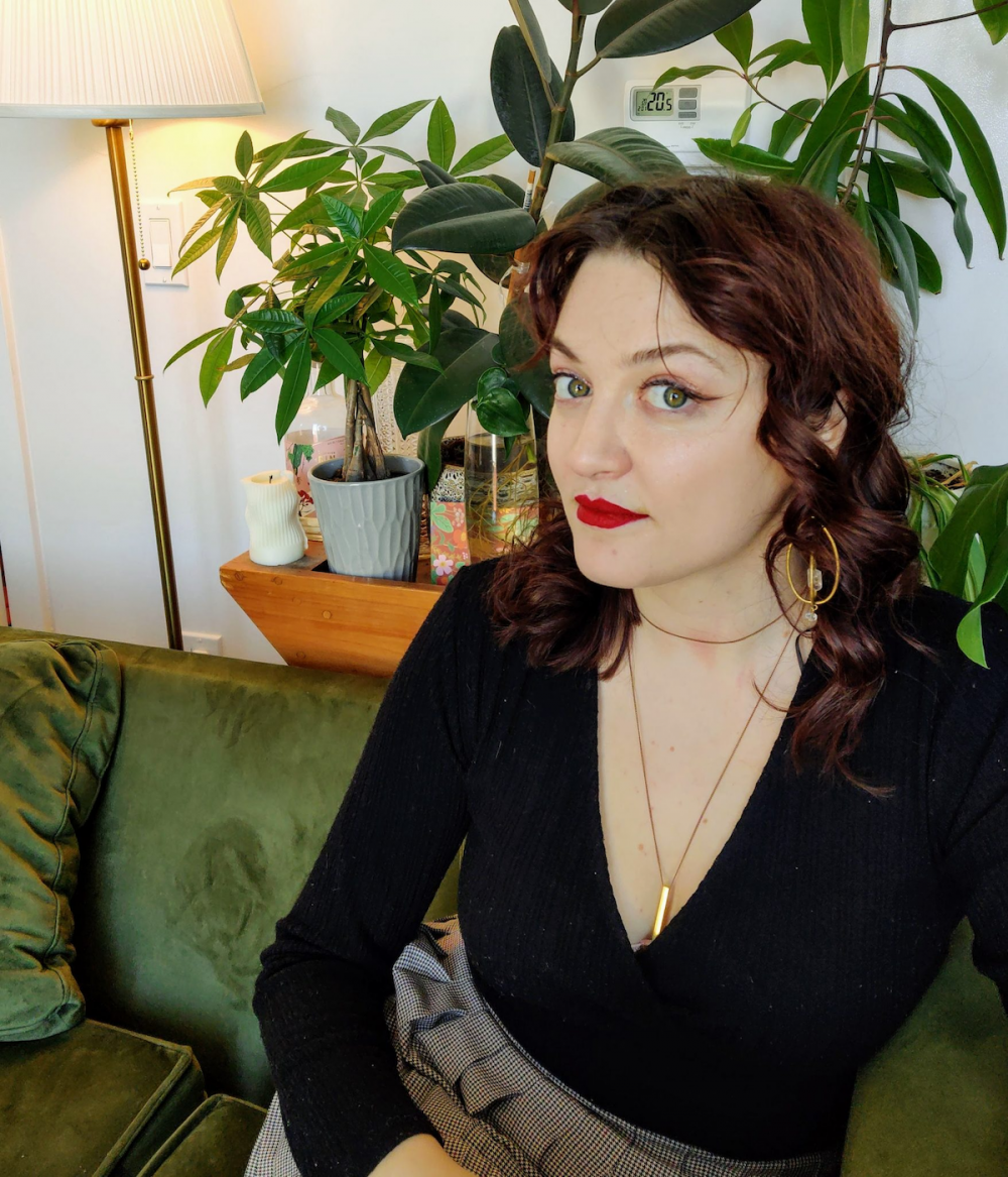 A disabled white femme with auburn hair and green eyes sits on a green velvet couch. She is wearing a black v-neck sweater and a pleated grey skirt. There are plants and beverages on a shelf behind her.