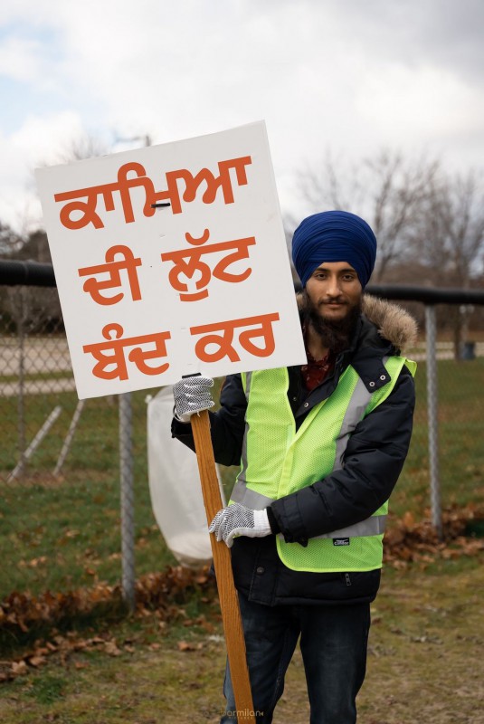 A photo of Navpreet holding a banner in Panjabi. The words on the banner translate to "Stop the exploitation of workers." He wears a yellow high-viz vest, a navy turban, and a slight smile.
