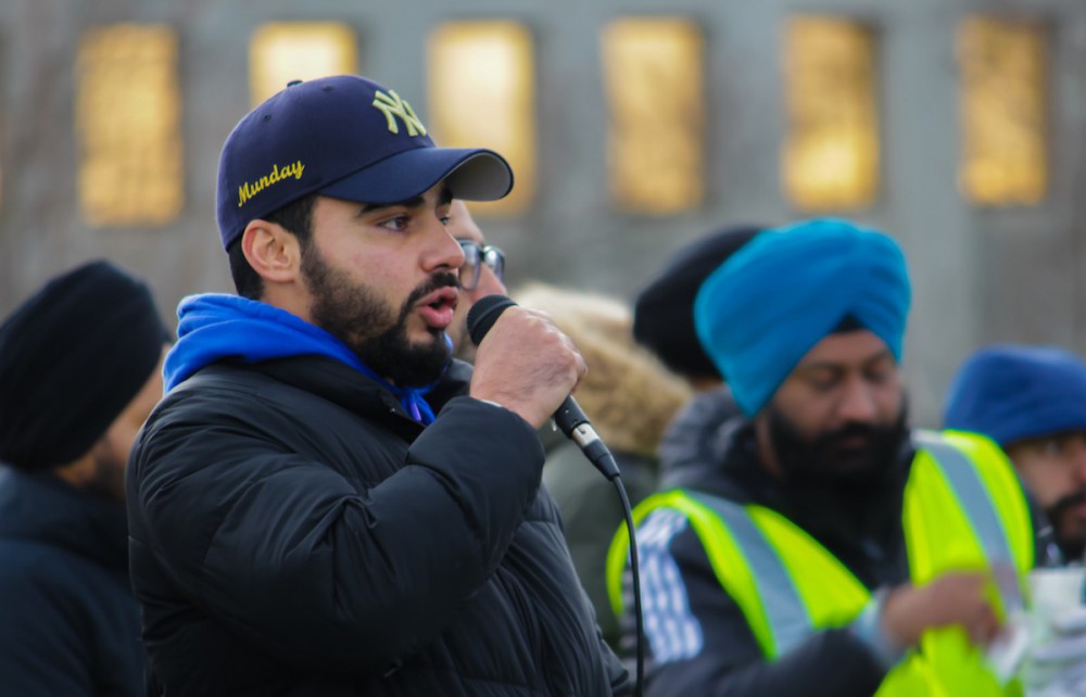 A close-up photo shows Parminder speaking into a microphone he's holding. Behind him, out of focus, are about 4 other men. Parminder has light brown skin, a short black beard, and is wearing a New York Yankees baseball cap.
