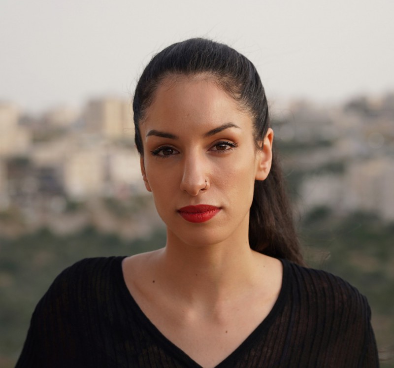 Rana Nazzal Hamadeh, a Palestinian woman, is standing in front of a blurred city landscape. She is wearing a black ribbed v-neck sweater and her long, black hair is tied back in a sleep ponytail. She has a small gold nose ring on her left nostril and she's wearing eyeline, mascara, and red lipstick.