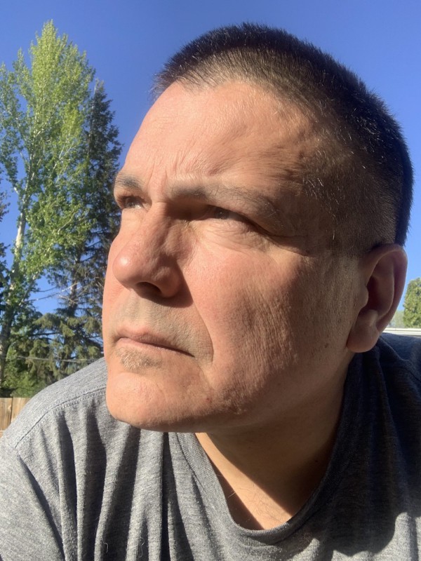Randy Lundy, a Cree man, is tilting his head to the right. He has short, black, recently buzzed hair, brown eyes, and a clean-shaven face. He is wearing a grey t-shirt. In the background are a clear blue sky, trees, and a wooden fence.