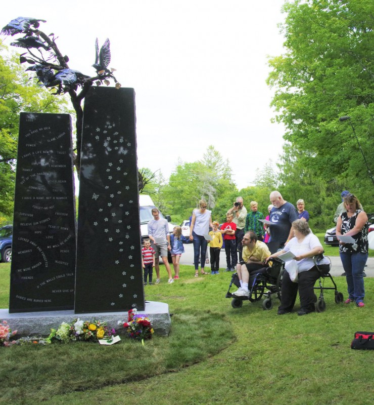 A photo of a large black stone monument, with words engraved on the front. It's standing on a green lawn and surrounded by a dozen people, including a person in a wheelchair, a number of children, and one person who is reading from a paper.