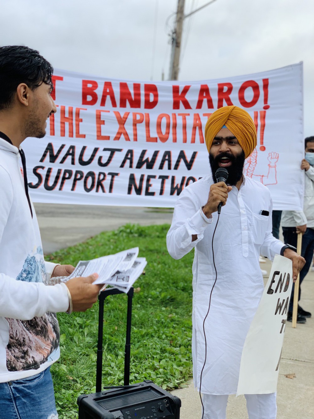 A person with a black beard, white salwar kameez, and orange turban speaks forcefully into a microphone in front of an NSN banner. To his left, partially out of frame, Rohit – who has short black hair and a white sweater – pauses to listen.