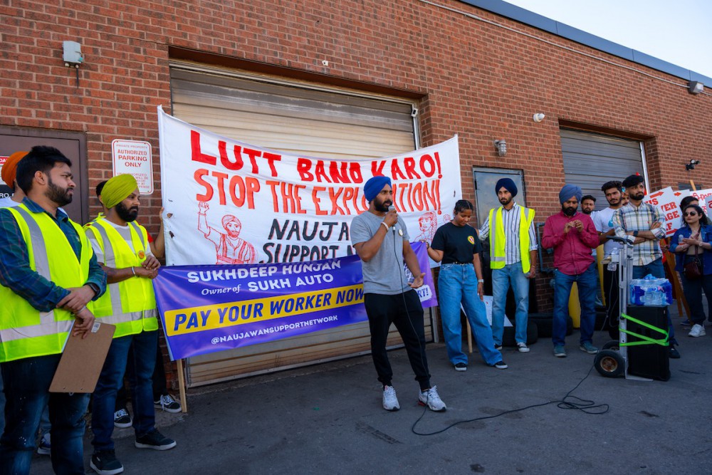 Rupinder stands in front of a brick building, wearing a blue turban and speaking into a microphone. He is surrounded by a dozen people wearing yellow high-viz vests. They are holding a banner that says