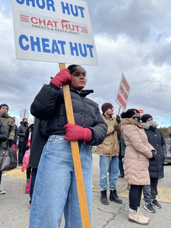 A photo of Satinder holding a sign that says "Chor Hut - Chat Hut - Cheat Hut." She has brown skin, a determined expression on her face, and is wearing jeans, a black puffer jacket, and red gloves.