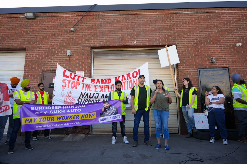 A woman with light brown skin speaks into a microphone while standing outside a garage. She is surrounded by a dozen people, most of whom are wearing yellow high-viz vests. They are holding a banner that says