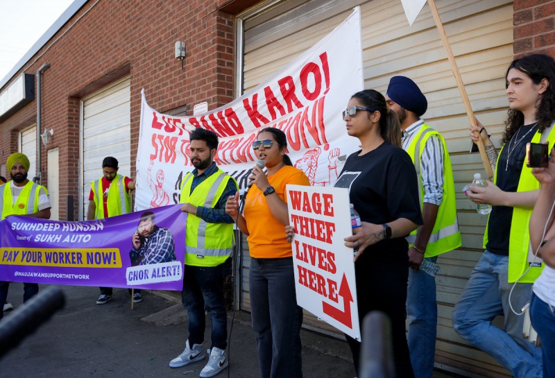 Simranjeet, a brown woman in an orange shirt and aviator sunglasses, holds a microphone and speaks in front of a brick building. She is surrounded by people holding signs saying "WAGE THIEF LIVES HERE" AND "SUKHDEEP HUNJAN, OWNER OF SUKH AUTO: PAY YOUR WORKER NOW!"