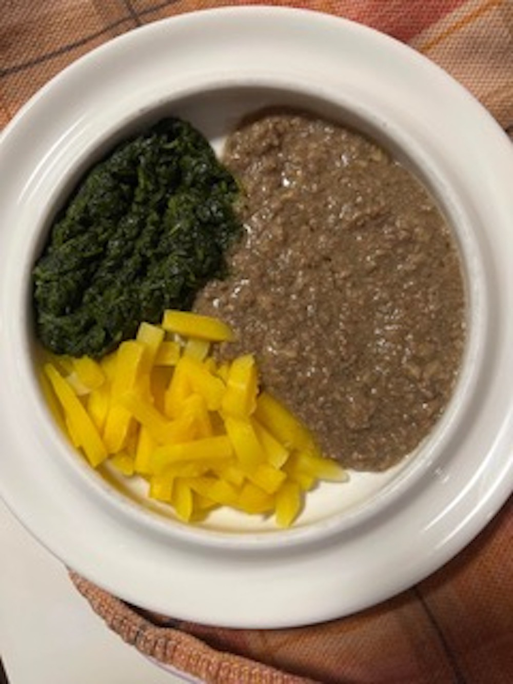 A photo of three types of food on a white plastic plate. The food is unidentifiable and consists of: yellow vegetable wedges, a dark green pulp, and a watery brown gruel.