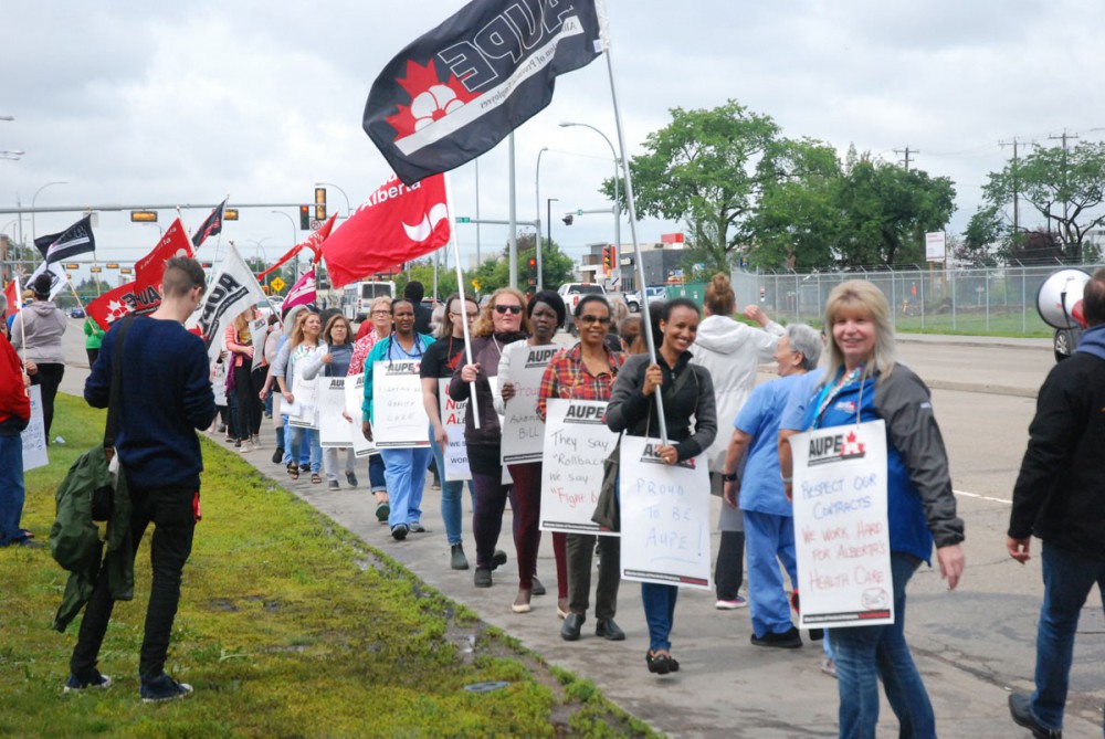 Nurses and allies walk a picket line on a sidewalk holding flags and signs denouncing Bill 9.