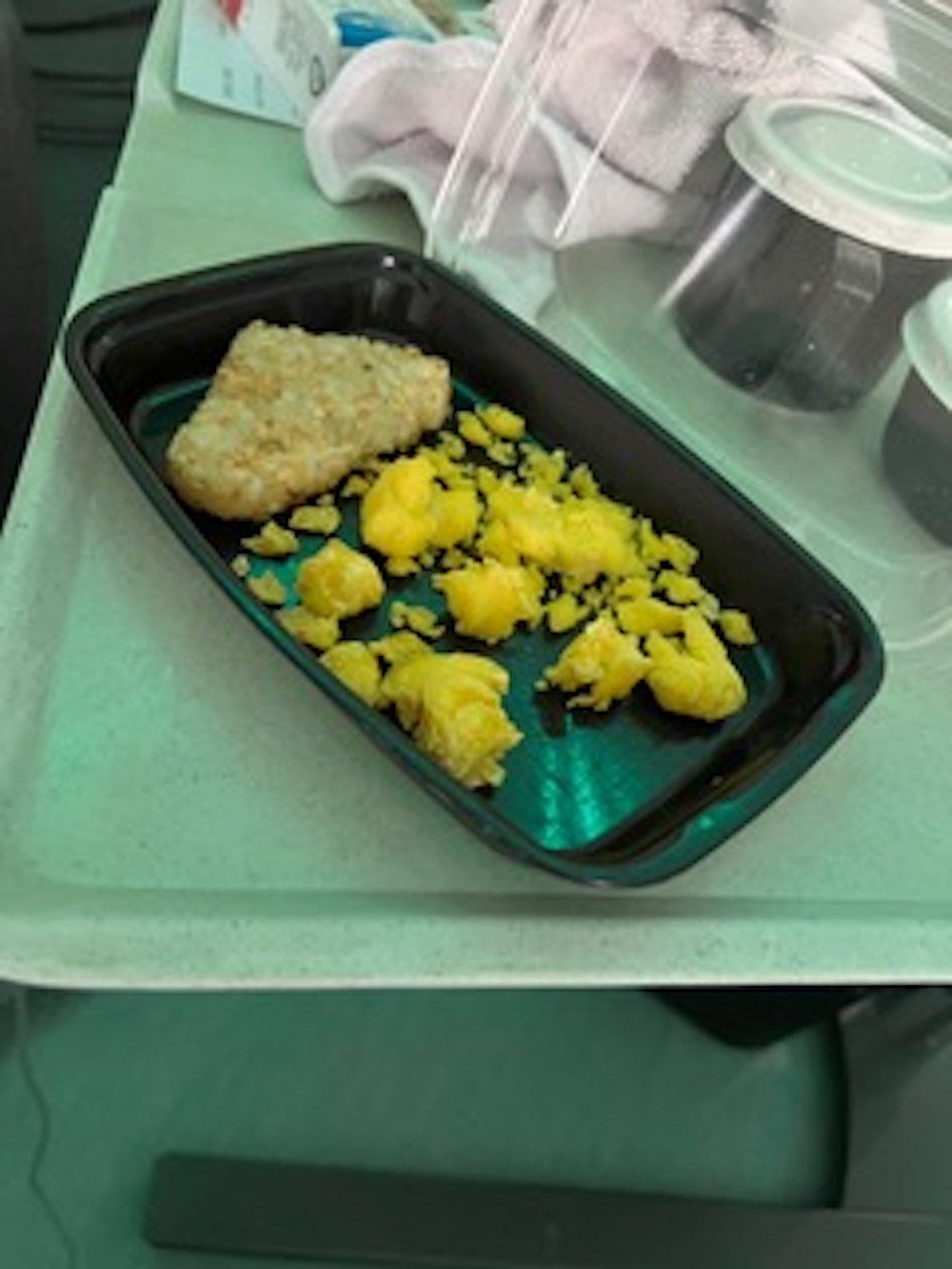 A photo of scrambled eggs and a hash brown in a black, disposable takeout container. The scrambled eggs do not fill the container – they are sparsely scattered across the bottom, and look very dry and chunky. The hash brown is pale.