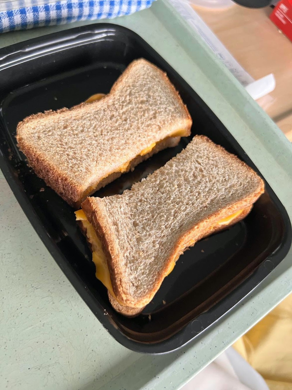 A photo of a cheese sandwich. The bread is plain and untoasted, and there is unmelted cheese visible between the two slices of bread. It is in a disposable black plastic container.