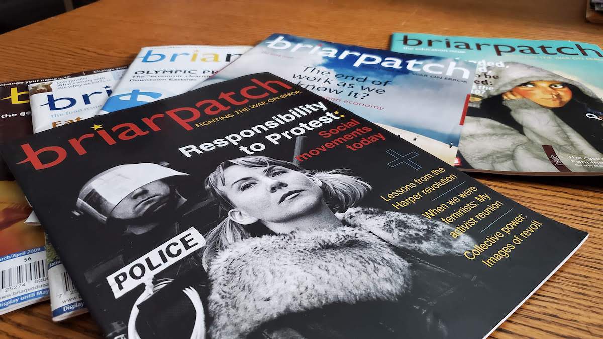 A photo of copies of Briarpatch from 2009 strewn across a wooden table. The cover of the topmost issue shows a police in riot gear looming behind a woman, with the words "Responsibility to protest: social movements today" and "Lessons from the Harper revolution."