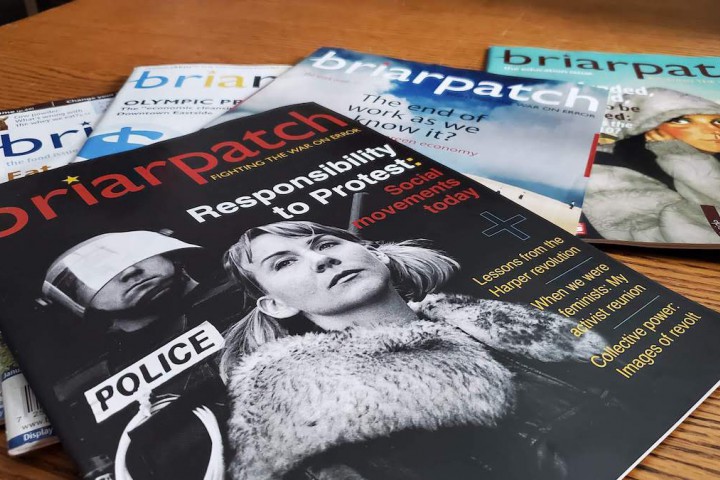 A photo of copies of Briarpatch from 2009 strewn across a wooden table. The cover of the topmost issue shows a police in riot gear looming behind a woman, with the words