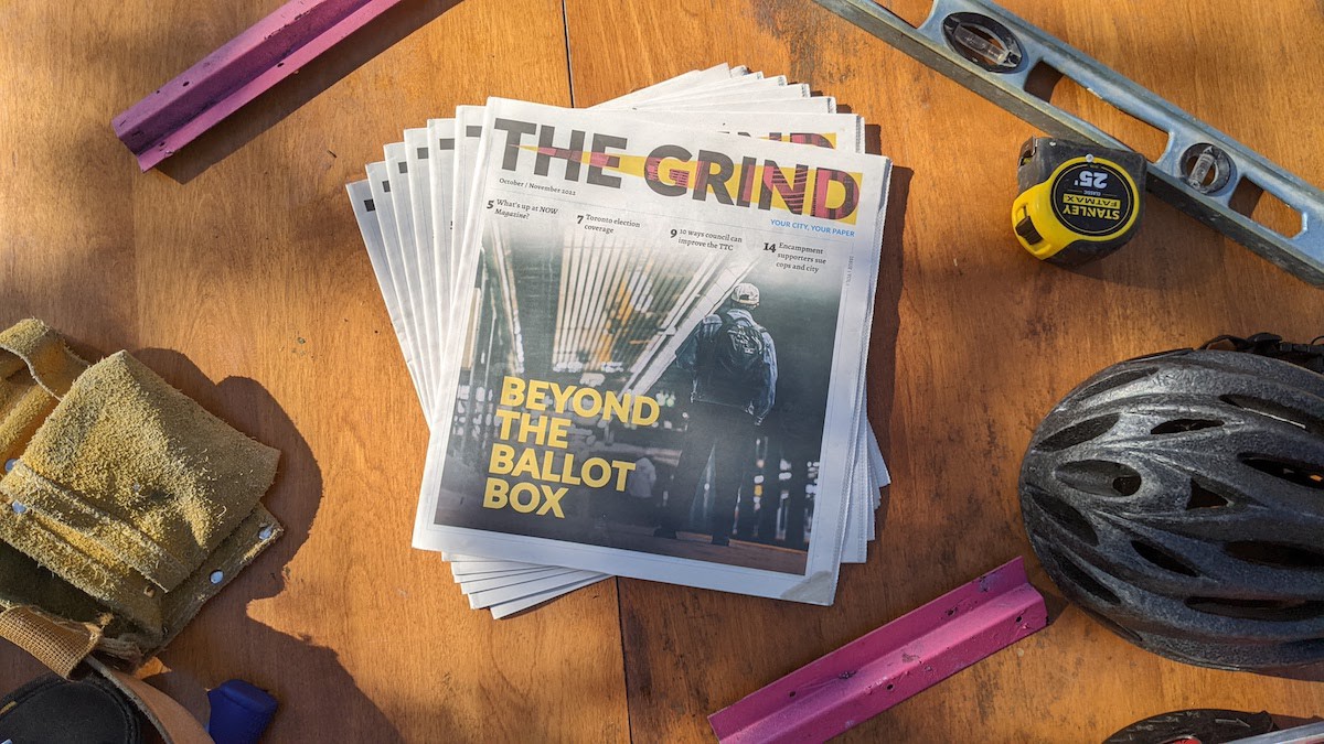 A photo taken from bird's-eye view. In the center, copies of an alt-weekly newspaper called The Grind are fanned out. The cover of The Grind shows a person standing in a subway station and the words "Beyond the Ballot Box." Around the newspaper various objects are scattered: a level, a measuring tape, a tool belt, a bike helmet.