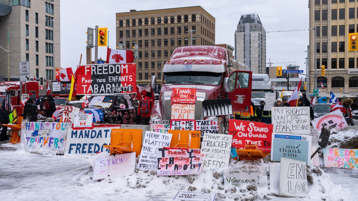 A photo of downtown Ottawa. Against a background of skyscrapers, huge trucks are parked, facing the camera. A multitude of signs lean up against them, with slogans like "Mandate Freedom" and "We Stand On Guard For Thee."