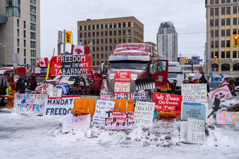 A photo of downtown Ottawa. Against a background of skyscrapers, huge trucks are parked, facing the camera. A multitude of signs lean up against them, with slogans like