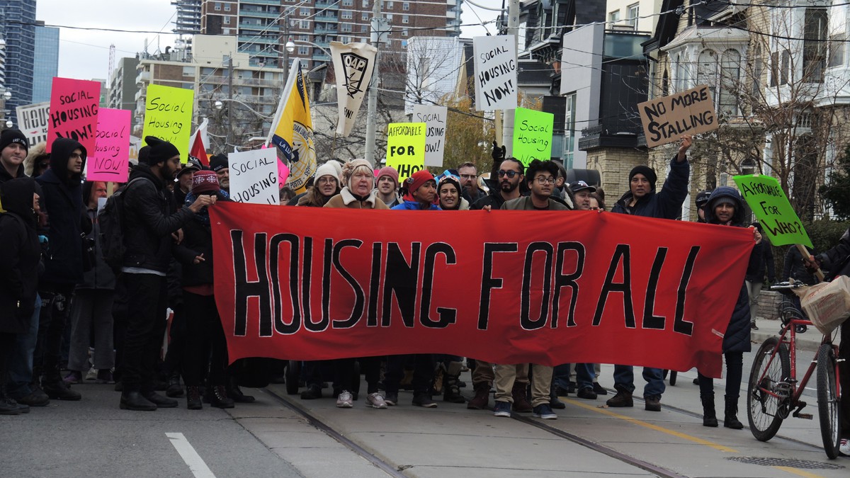 Ani-poverty activists marching in Toronto holding colourful banners, including a large red banner that says "HOUSING FOR ALL."