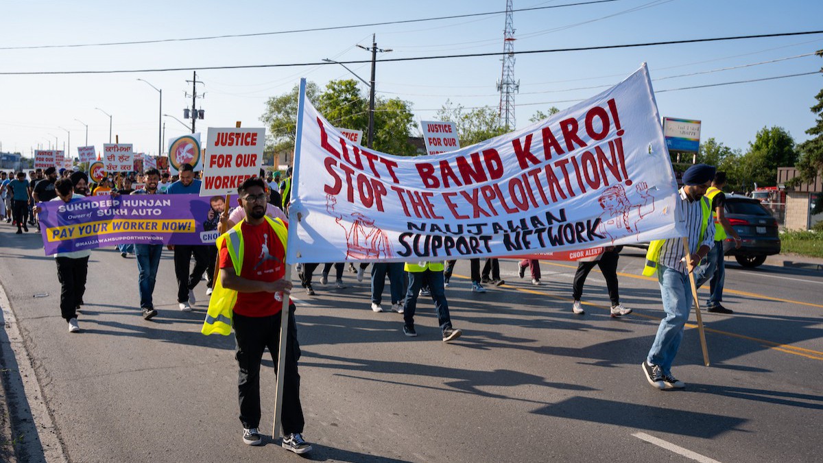 A photo of people marching on a road in Brampton. Two people in the front are carrying a large banner that says "LUTT BAND KARO / STOP THE EXPLOITATION / NAUJAWAN SUPPORT NETWORK." Behind them, dozens of others carry signs in Panjabi.