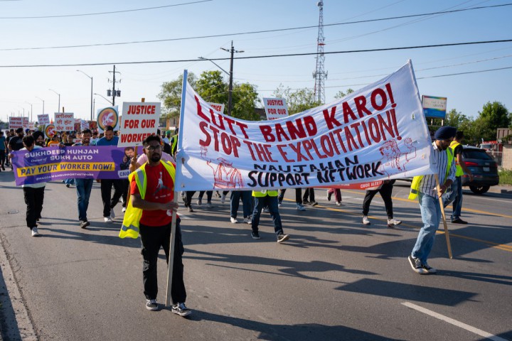 A photo of people marching on a road in Brampton. Two people in the front are carrying a large banner that says