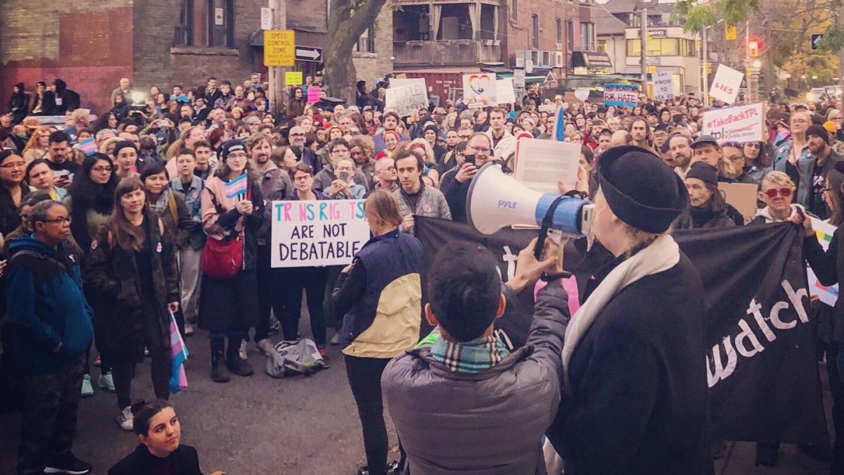 A photography of a crowd from above, listening to someone speak through a megaphone. One member of the crowd holds a sign that says "Trans rights are not debatable."