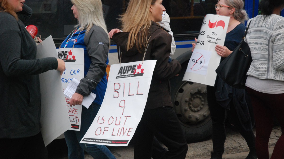 A person with long hair walks a picket line among others, wearing a sign that says