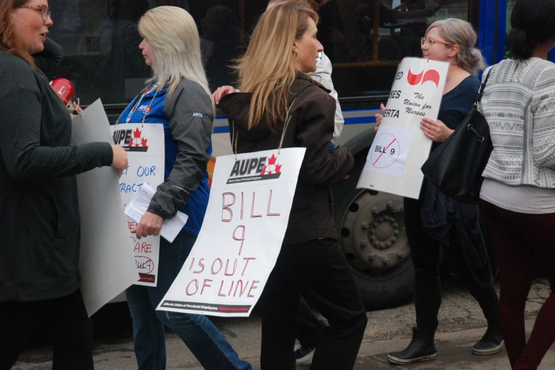 A person with long hair walks a picket line among others, wearing a sign that says
