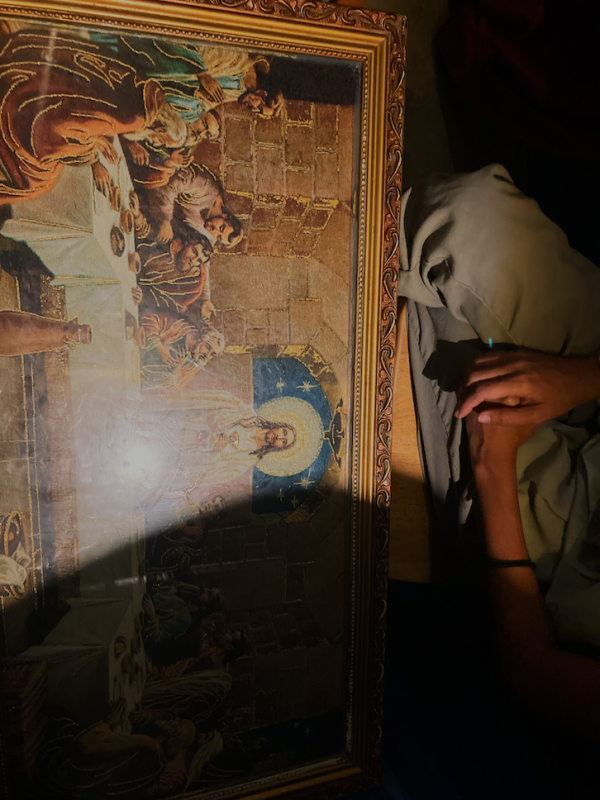 Filipina photographer Hannah Reyes Morales kneels by a painting of a the Last Supper. Only her knees and crossed hands are visible above the top two thirds of the painting.